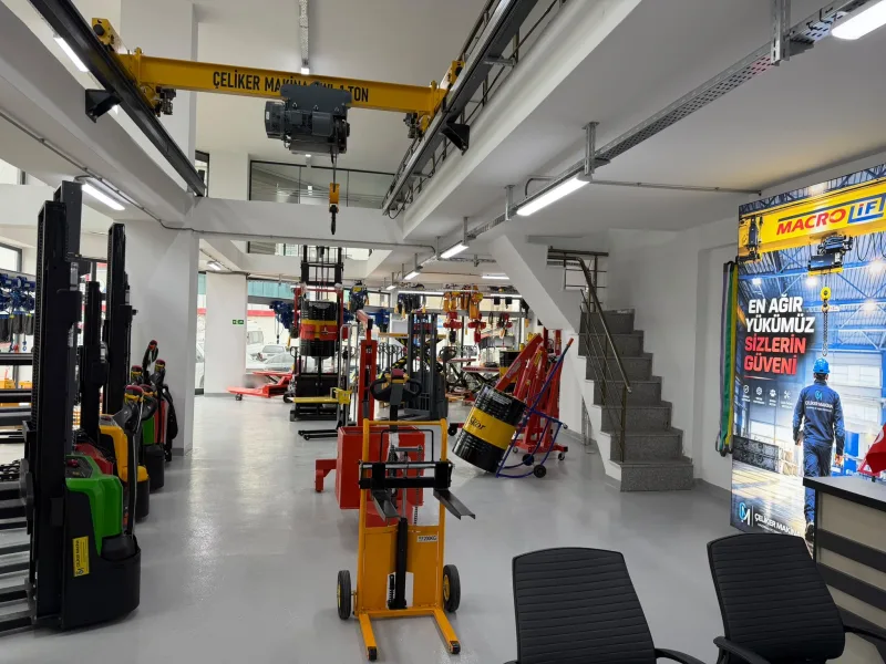 A yellow single girder 1 t overhead crane with an electric wire rope hoist on the showroom ceiling, with stackers and a drum trolley below