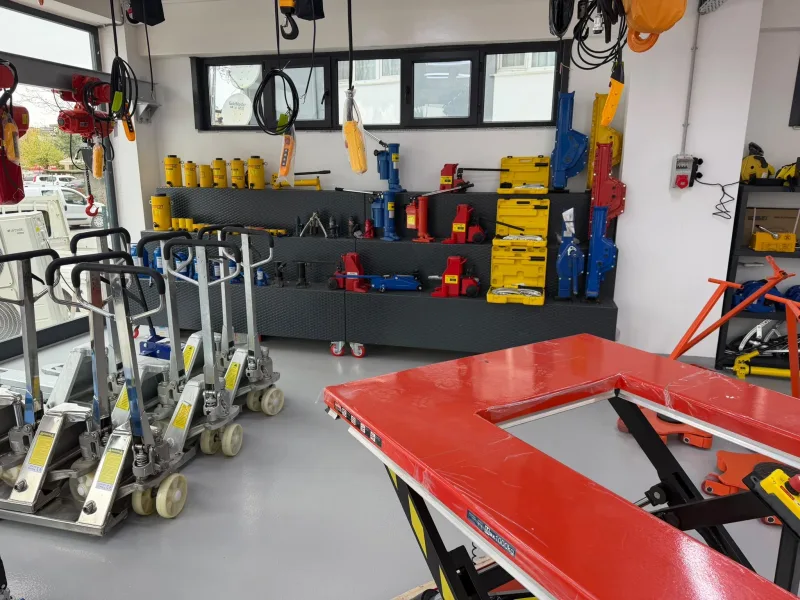 Yellow hydraulic cylinders and a hand pump, red and blue hydraulic jacks and mechanical jacks on display, with galvanised pallet trucks and a U-shaped lift table in front
