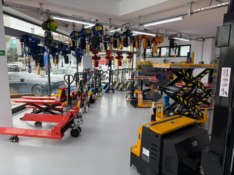 Blue, red and orange Macrolift electric chain hoists hanging from ceiling beams in the showroom, with hand pallet trucks and a scissor lift table below