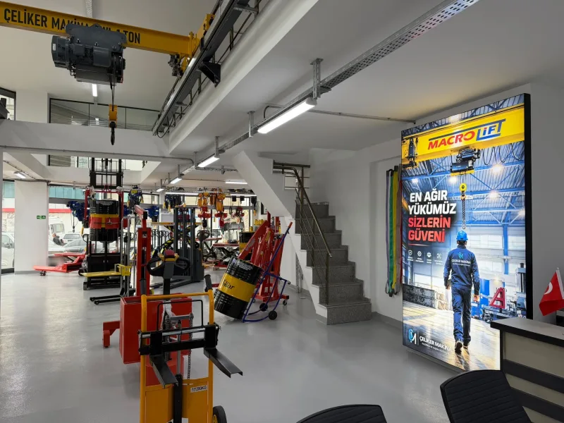 Inside the showroom: a 1 t overhead crane on the ceiling, chain hoists hanging at the back, a manual stacker and a drum trolley in front and a Macrolift display panel on the right
