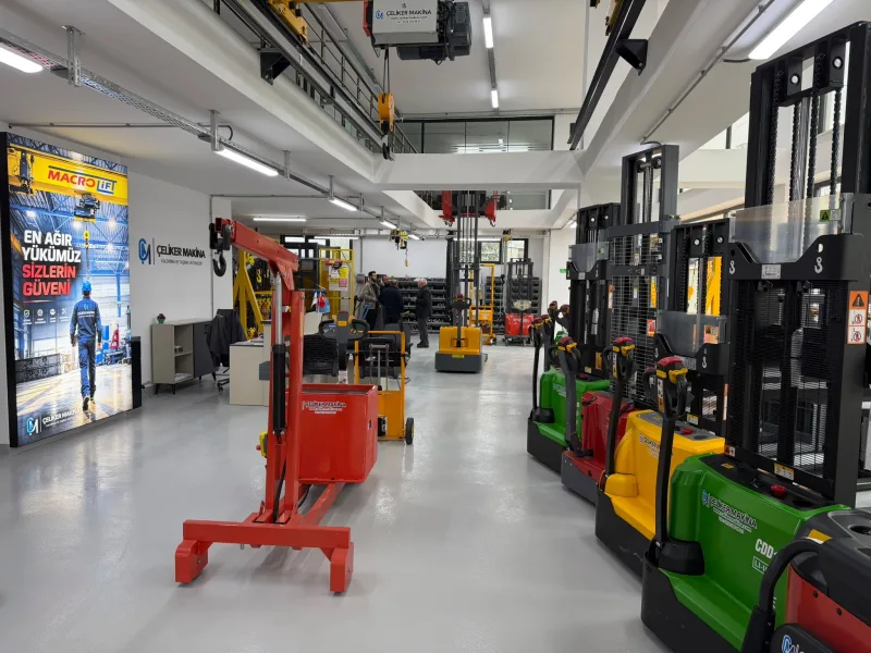 A red mobile workshop crane with green, red and yellow electric stackers on the right and the Çeliker Makina wall sign and Macrolift panel at the back