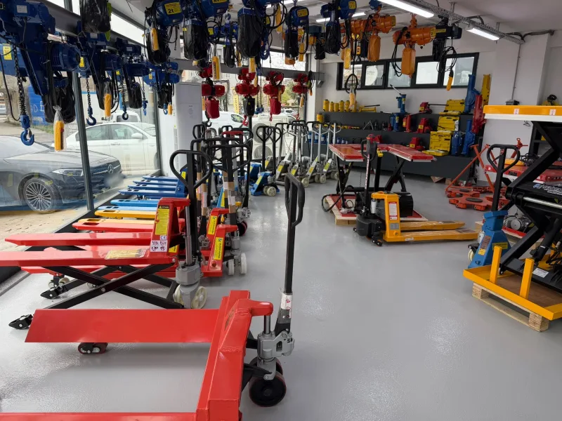 Red, blue and galvanised hand pallet trucks and a scissor pallet truck by the shop window, with a hydraulic jack display, lift tables and chain hoists behind